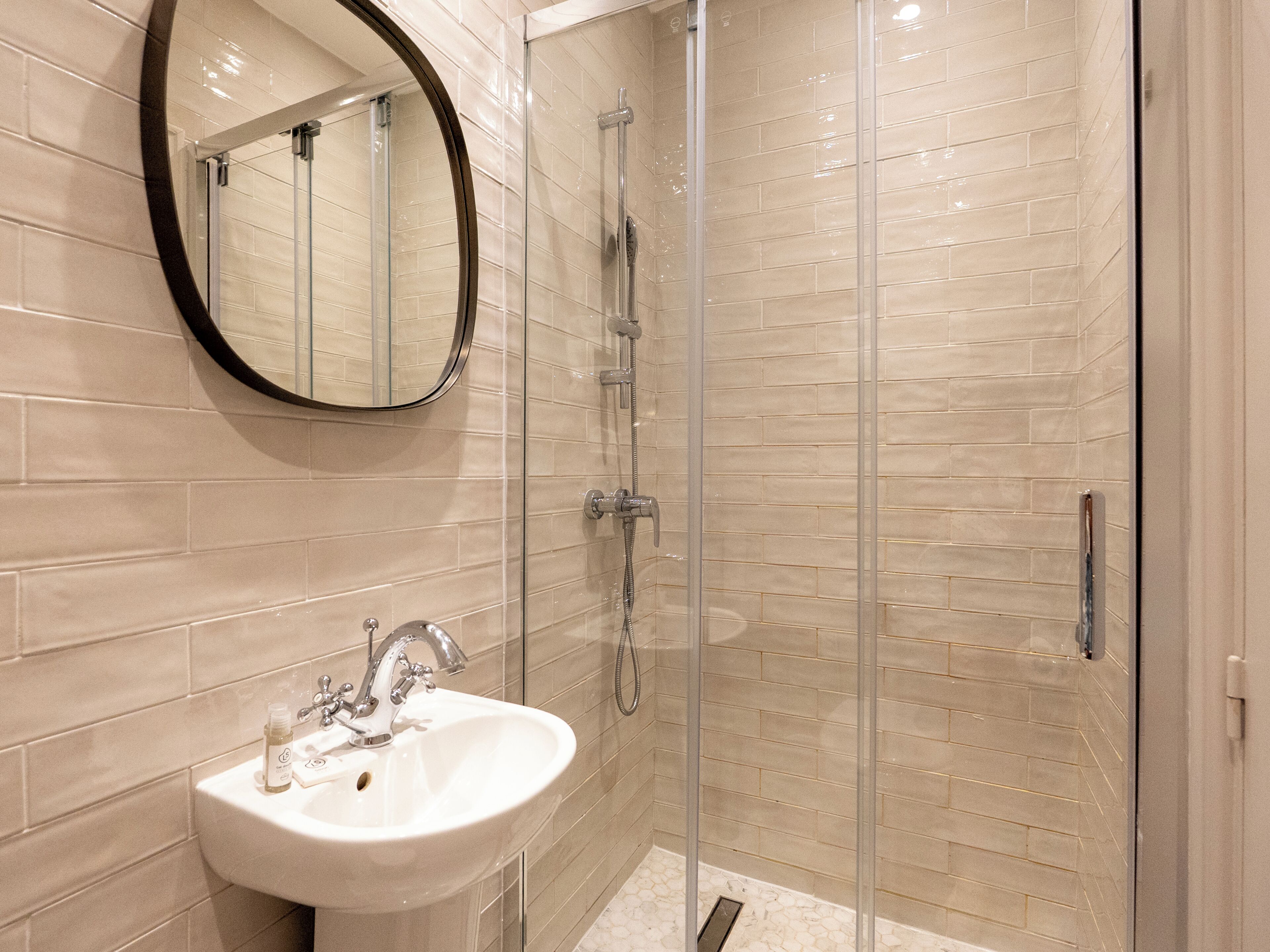 Une salle de bain avec une douche à parois de verre, un lavabo blanc avec un miroir au-dessus, et des murs recouverts de carreaux beiges.