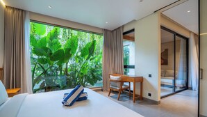 Une chambre dotée d’un grand lit, d’un bureau en bois et d’une porte-fenêtre coulissante donnant sur un balcon avec vue sur une végétation luxuriante.
