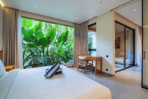 A bedroom with a large bed, a wooden desk, and a sliding glass door leading to a balcony with a view of lush greenery. - Novo Ubud by Betterplace (Ubud)