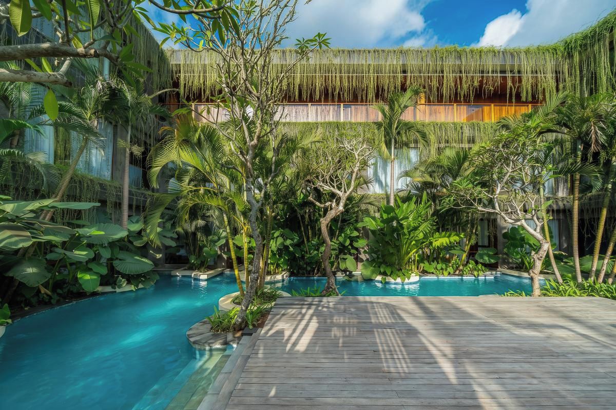 Área de piscina com deck de madeira, cercada por plantas tropicais e um edifício com telhado de palha.
