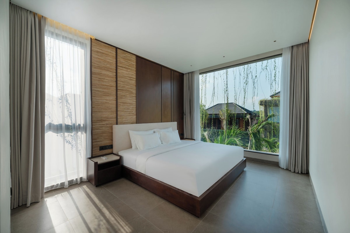 Habitación de hotel moderna con una cama grande, una mesita de noche, un panel de pared de madera y vistas a zonas verdes por la ventana.