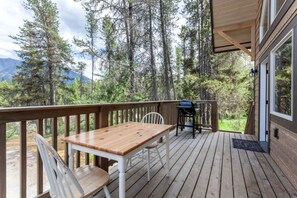 Een houten terras met een tafel, stoelen, een barbecue en uitzicht op een bos en bergen.