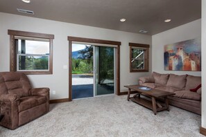 A living room with a brown leather sofa, a wooden coffee table, and a beige carpet. There are large windows with wooden frames, a sliding glass door, and a painting on the wall. - Family-Friendly Cabin Rental with Mountain Vistas near West Glacier, Montana (West Glacier)