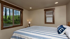 A bedroom with a large bed, a window with a view of trees, a bedside table with a lamp, and a framed picture on the wall. - Family-Friendly Cabin Rental with Mountain Vistas near West Glacier, Montana (West Glacier)
