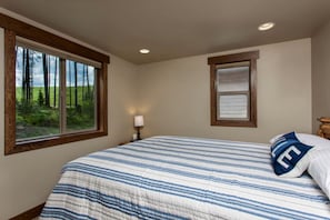 Un dormitorio con una cama grande, una ventana con vistas a árboles, una mesita de noche con una lámpara y un cuadro enmarcado en la pared.