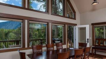 A dining area with a wooden table and chairs, large windows with a view of trees and mountains, and a hanging light fixture.