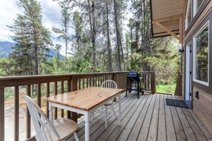 Eine Holzterrasse mit Tisch, Stühlen, einem Grill und Blick auf Wald und Berge.