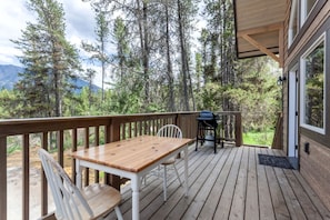 Una terraza de madera con una mesa, sillas, una parrilla y vistas a un bosque y montañas.