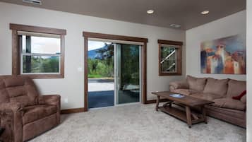 Ein Wohnzimmer mit braunem Ledersofa, einem Holztisch und einem beigefarbenen Teppich. Große Fenster mit Holzrahmen, eine Glastür und ein Gemälde an der Wand sind zu sehen.