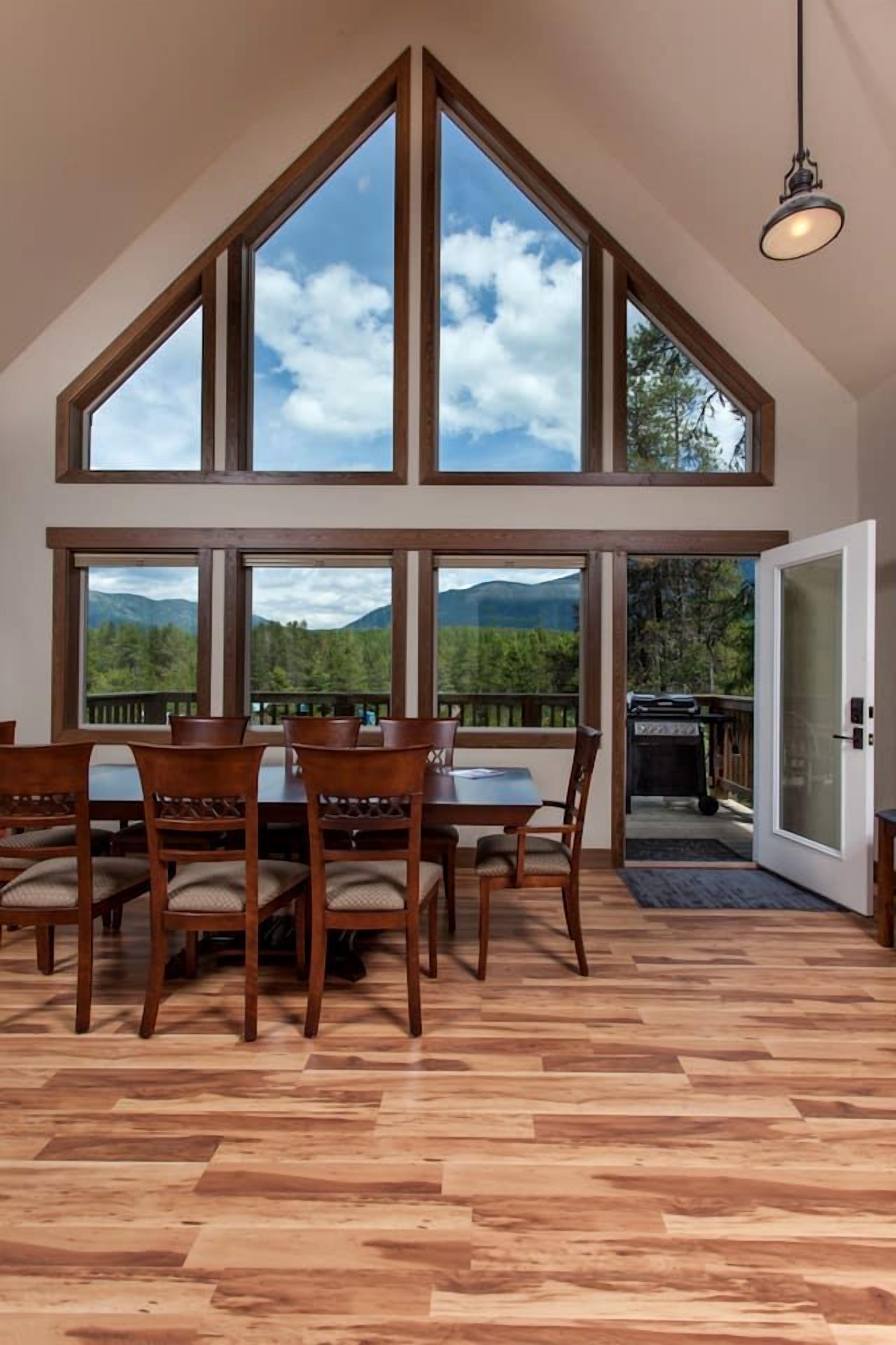 A dining area with a high ceiling and large windows, wooden flooring, and a view of mountains.