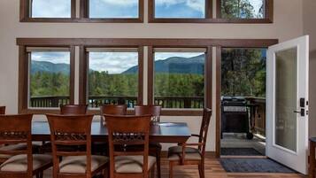 A dining area with a high ceiling and large windows, wooden flooring, and a view of mountains.