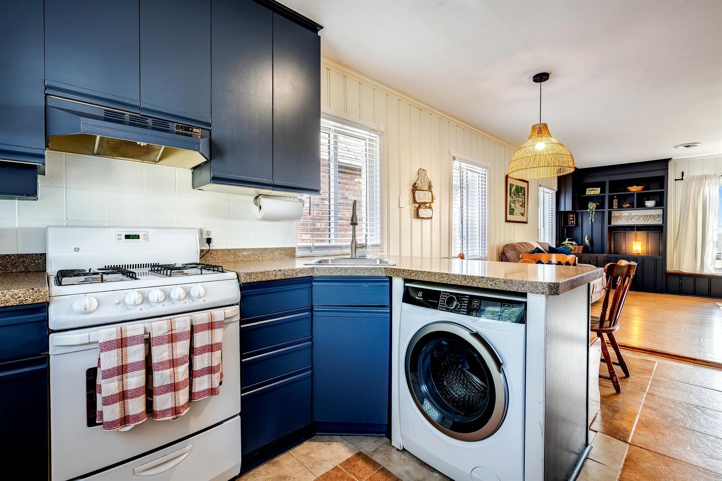 A compact kitchen with a stove, oven, and washing machine, a small island with seating, and a dining area with a chandelier.
