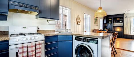 A compact kitchen with a stove, oven, and washing machine, a small island with seating, and a dining area with a chandelier.