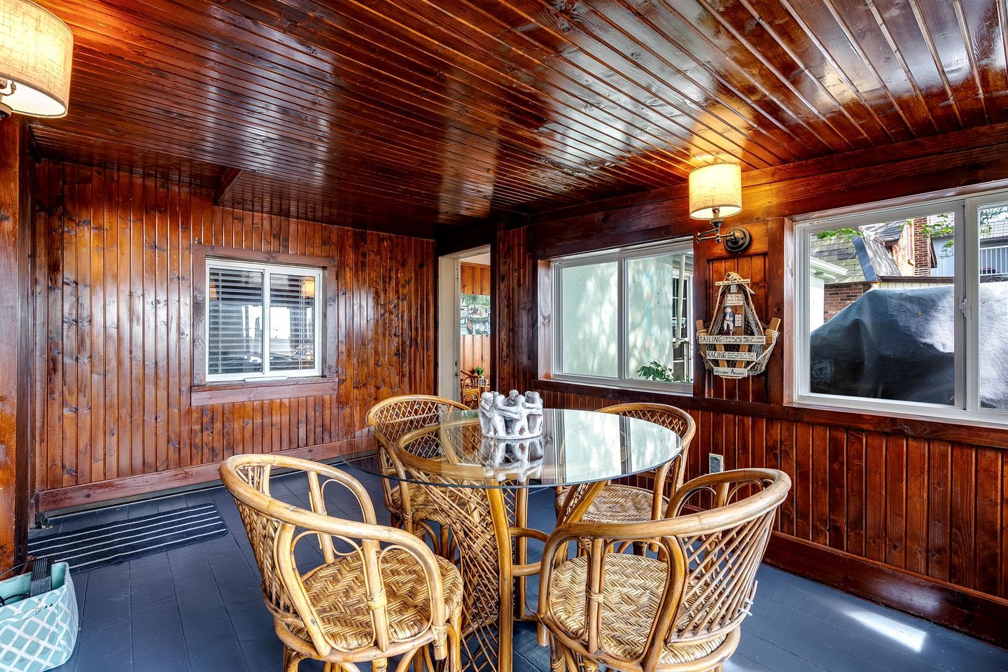 Una habitación con paneles de madera, una mesa redonda de vidrio y sillas de mimbre, una ventana con persianas y una lámpara fijada a la pared.