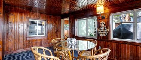 Una habitación con paneles de madera, una mesa redonda de vidrio y sillas de mimbre, una ventana con persianas y una lámpara fijada a la pared.