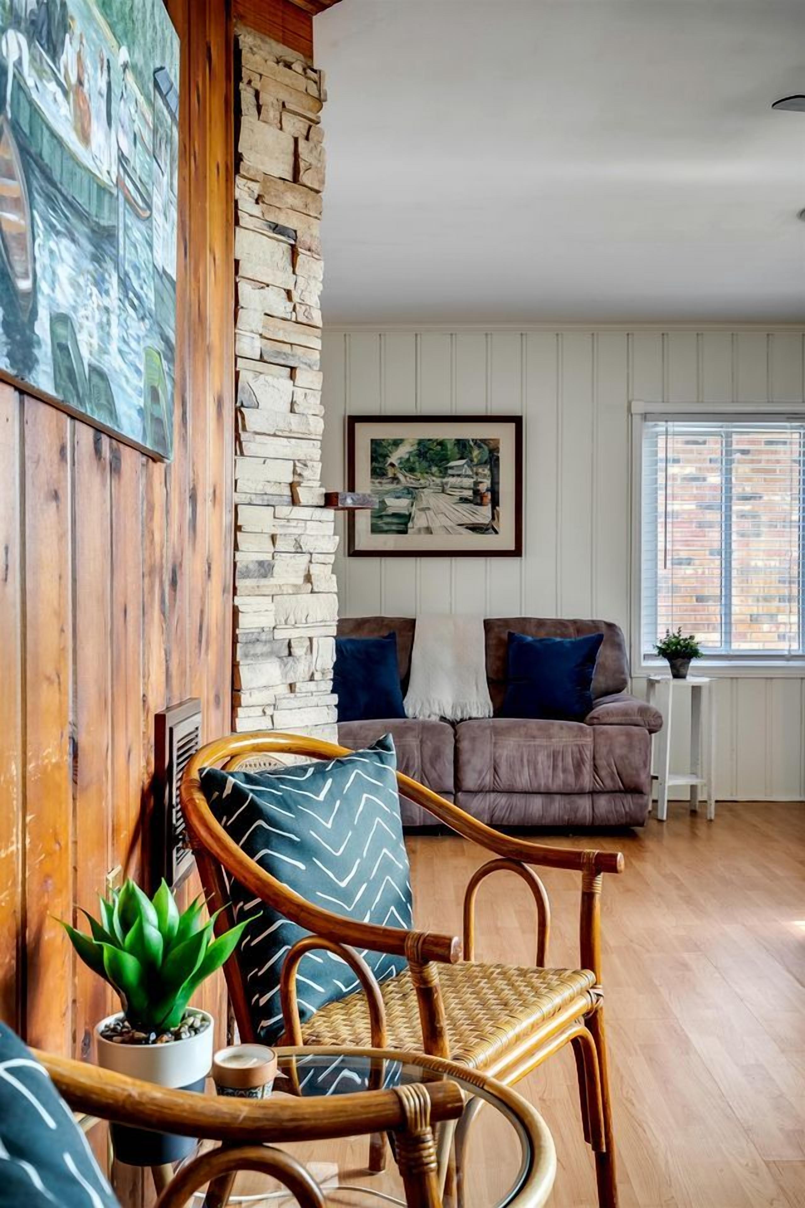 Una sala de estar acogedora con pared de piedra, techo de madera, un sofá marrón, un sillón de mimbre y una pequeña mesa con una planta.
