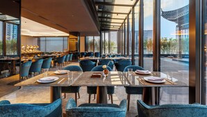 En moderne restaurant med høje lofter, store vinduer og udsigt til en traditionel bygning. Bordene er dækket med tallerkener og glas, og der er blå stole.