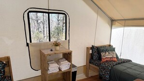 Interior acolhedor de uma RV com prateleira de madeira com toalhas, janela com vista para árvores e cama com manta estampada e almofada com frase motivacional.