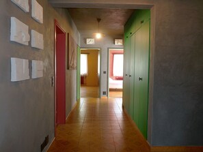 Un pasillo con piso de baldosas, una puerta roja y una puerta verde. Hay interruptores de luz en la pared y un accesorio de iluminación en el techo.
