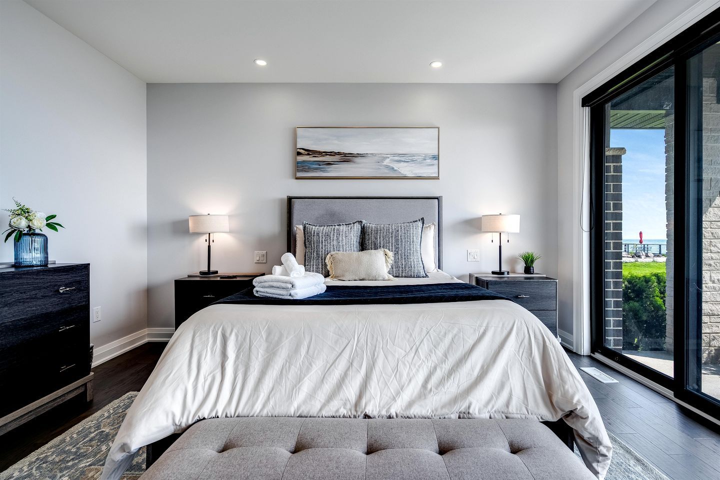 A modern bedroom with a large bed, two bedside tables with lamps, a view of the outdoors through a large window, and a painting above the bed.