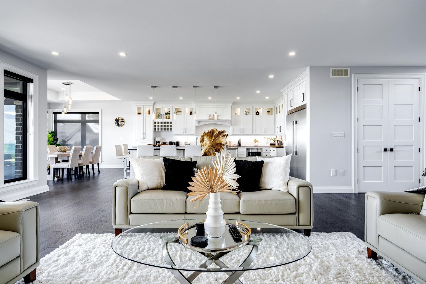 A modern living room with a glass coffee table, a beige sofa, and a dining area with white cabinets.
