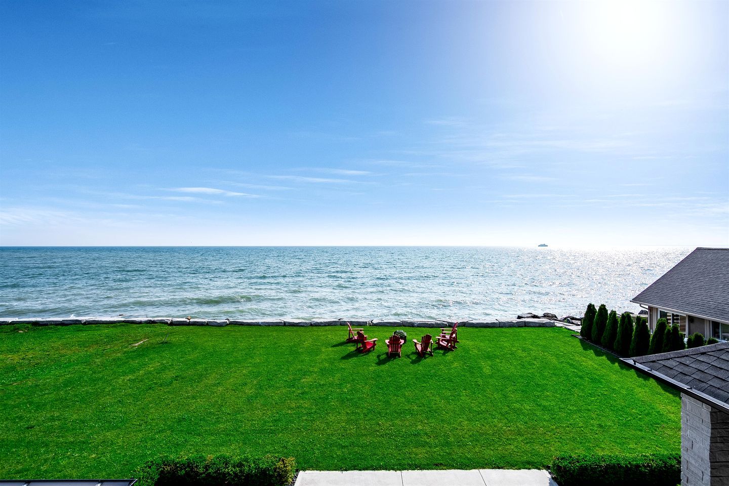 A grassy lawn with a view of the sea, a building with a sloped roof, and a few red chairs.