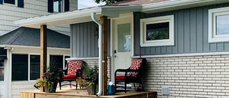 A house with a covered porch, two chairs, a door, and a brick exterior.