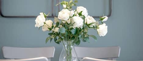 A dining table with a vase of white flowers, two white chairs, and a wall clock.