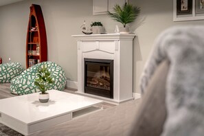 Un salon avec une cheminée blanche, des sièges à motifs verts, une table basse blanche et une bibliothèque remplie de livres et d’objets décoratifs.