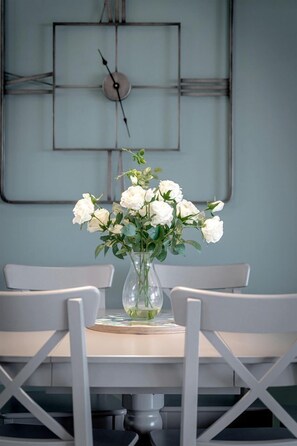 A dining table with a vase of white flowers, two white chairs, and a wall clock.