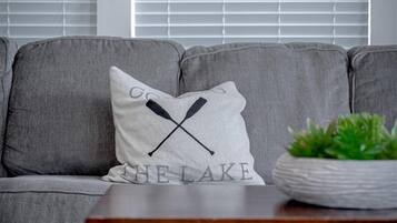 A grey sofa with a pillow featuring a black X and the text 'THE LAKE.' A wooden table with folded blankets is in front of the sofa.