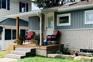 A house with a covered porch, two chairs, a door, and a brick exterior.