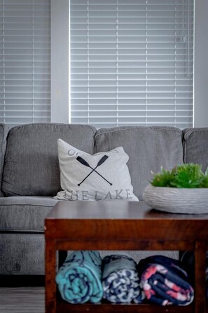 A grey sofa with a pillow featuring a black X and the text 'THE LAKE.' A wooden table with folded blankets is in front of the sofa.