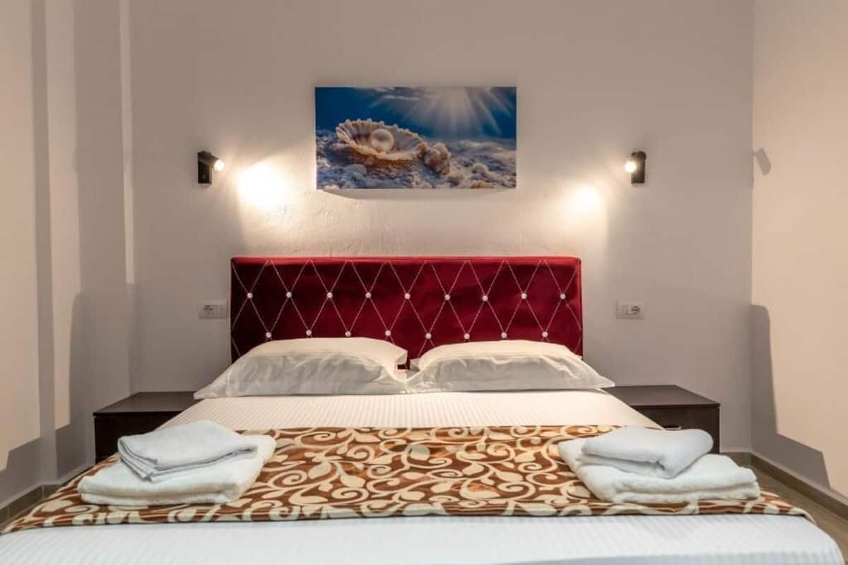 Habitación de hotel con cama, dos almohadas, cabecero rojo, un cuadro colgado en la pared y dos mesitas de noche.