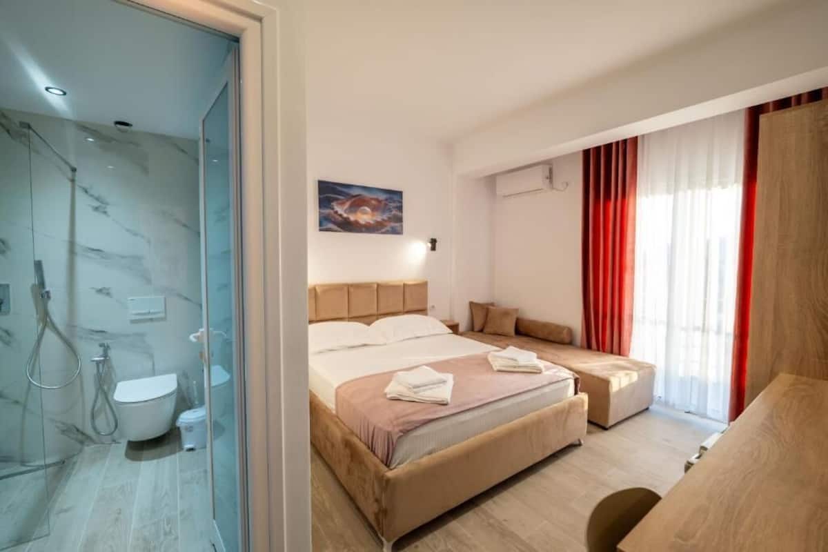 Una habitación de hotel moderna con cabina de ducha de vidrio, cama con cabecero, mesita de noche y ventana con cortinas rojas.