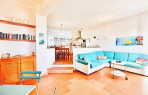 A living room with a blue sofa, a wooden cabinet, and a dining area with chairs. - Hostdomus - In the Blue - Bike and Sea (Finale Ligure)
