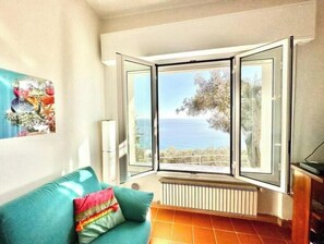 A room with a teal sofa, a radiator, and a bay window with a view of the sea. - Hostdomus - In the Blue - Bike and Sea (Finale Ligure)