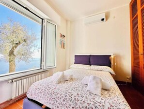 A bedroom with a large window, a bed with white linens, a radiator, and a wooden door. - Hostdomus - In the Blue - Bike and Sea (Finale Ligure)