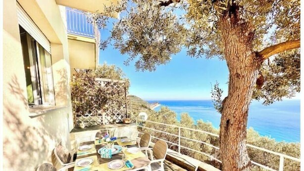 Area pranzo all'aperto con tavolo apparecchiato per quattro, circondato da sedie bianche, con vista sulla costa.