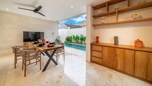 A modern living room with a dining area, a TV, a ceiling fan, a pool, and a wooden cabinet with decorative items. - Lekatika Villa (Kerobokan)