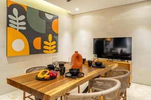 A dining area with a wooden table, wicker chairs, a TV, and a colorful abstract painting on the wall. - Lekatika Villa (Kerobokan)