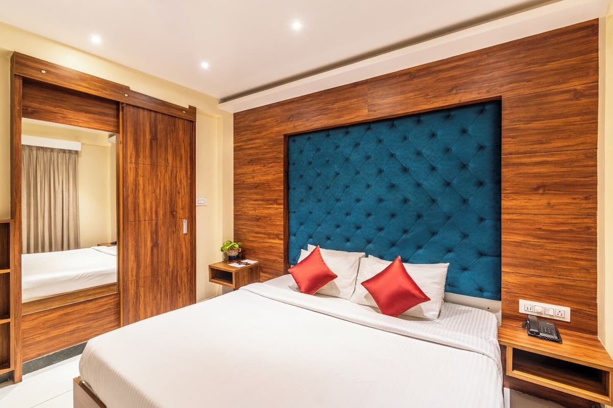 A hotel room with a large bed, wooden wardrobe, a blue upholstered headboard, and a bedside table with a lamp.