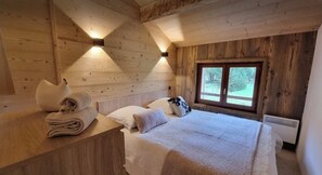 Habitació d'una cabana de fusta amb un llit, almoades, una tauleta de nit i una finestra.