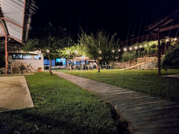 Espacio exterior nocturno con un sendero empedrado, asientos y un edificio con ventanas iluminadas.