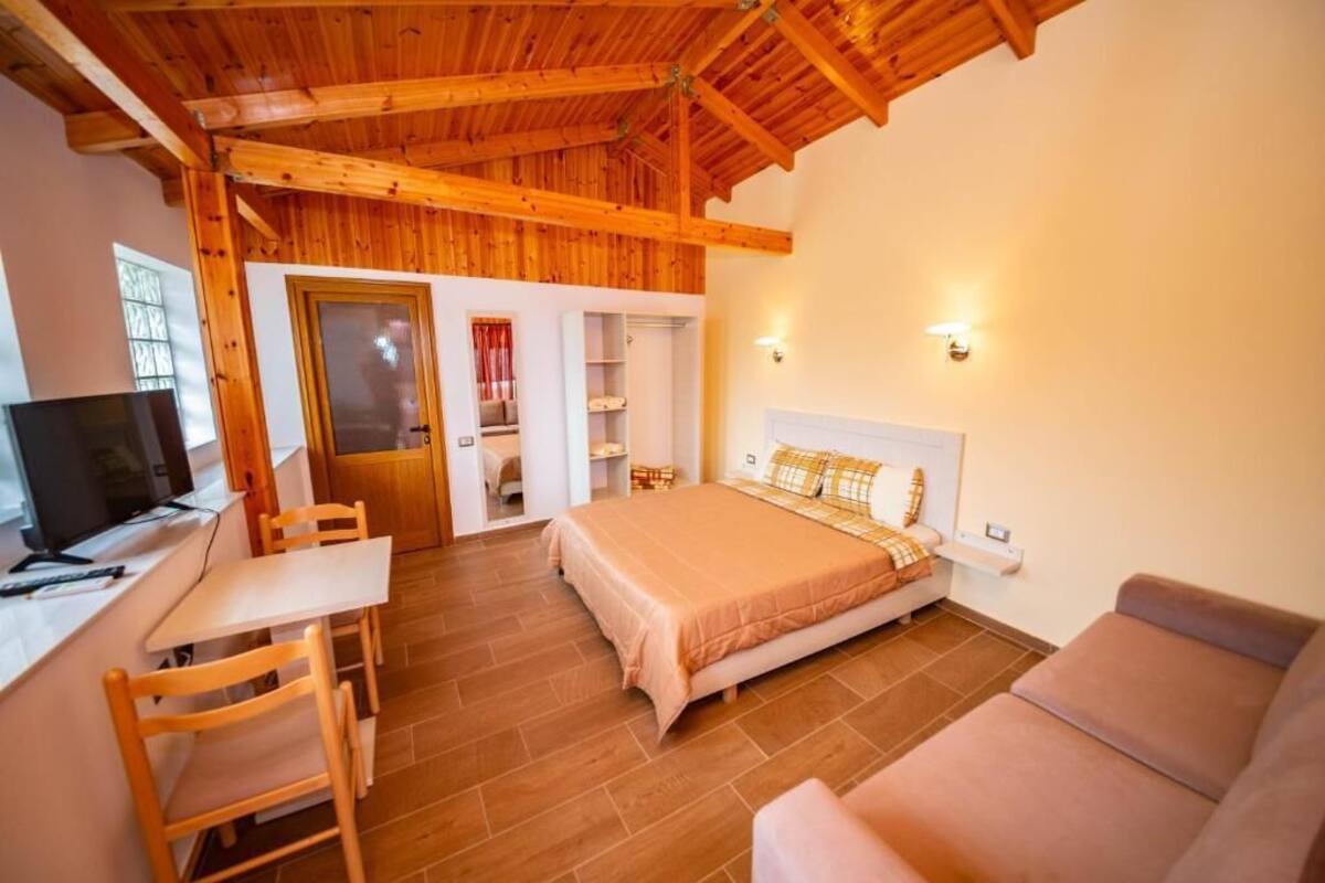 A bedroom with a wooden ceiling, a bed with a beige bedspread, a desk with a chair, and a television.