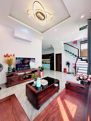 A modern living room with a flat-screen TV, wooden furniture, a staircase, and a ceiling light fixture. - Căn Hộ Ngay Trung Tâm Thành Phố Huế Đầy Đủ tiện Nghi, sân vườn rộng thoáng (Thành phố Huế)