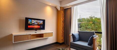 A modern living room with a flat-screen TV, a sofa, a coffee table, and a view of greenery outside.