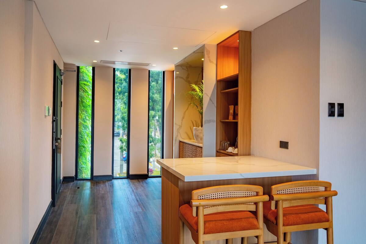 Un interior moderno con piso de madera, una zona de cocina con cubierta blanca y sillas de madera, y vista a follaje a través de la puerta.