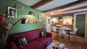 A cozy living room with a red sofa, a dining table with chairs, a kitchen in the background, and a wooden ceiling. - Studio Le Mélèze (Méolans-Revel)
