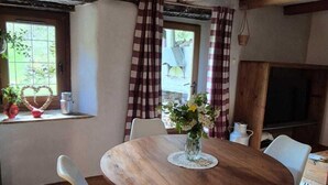 Un comedor con una mesa redonda de madera, sillas blancas, un florero con flores y una ventana con cortinas.